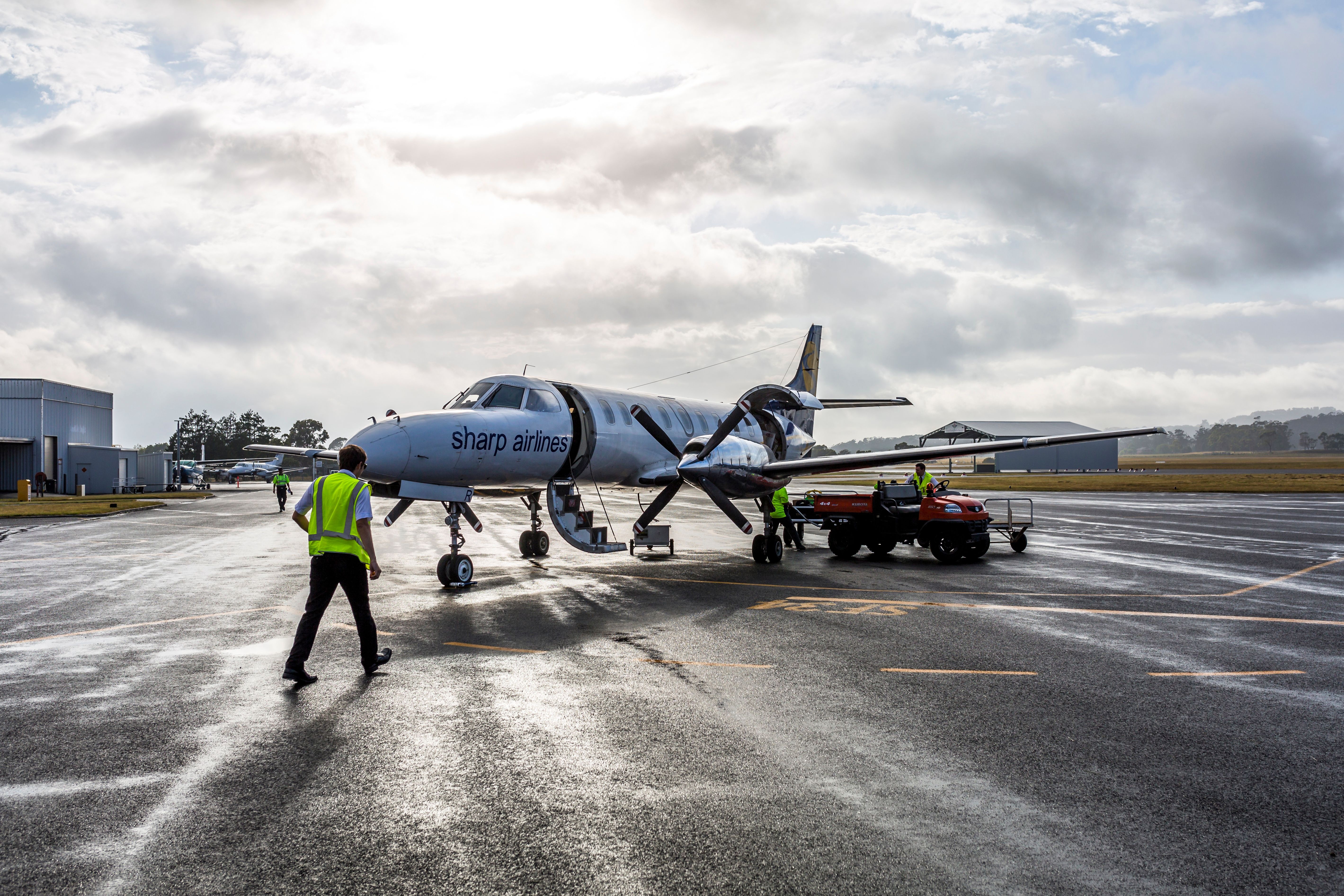 Ground crew in high visibility vests working around a Sharp Airlines propeller aircraft on a wet tarmac, King Island Airport