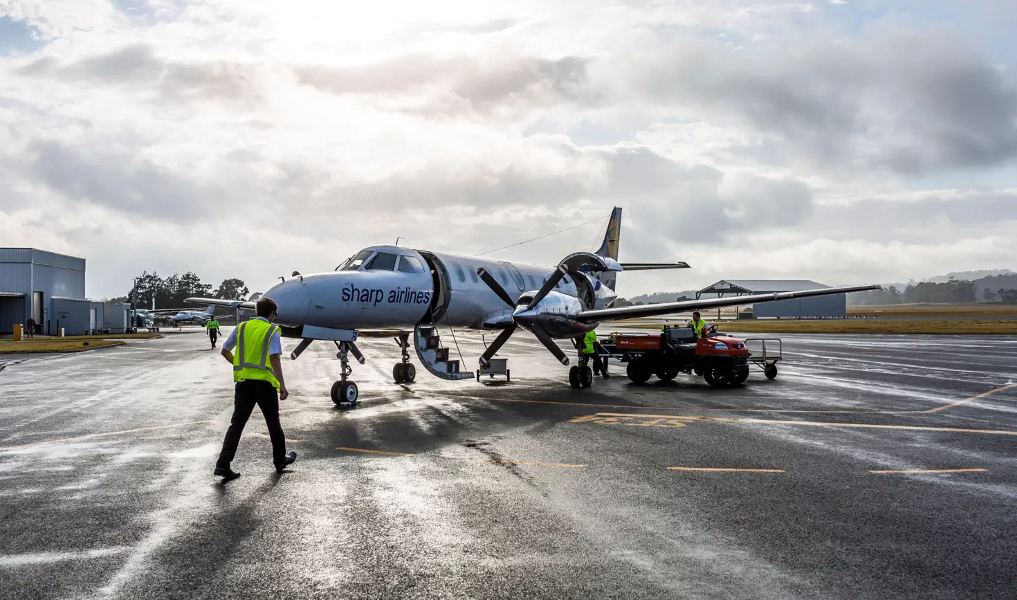 Ground crew in high visibility vests working around a Sharp Airlines propeller aircraft on a wet tarmac, King Island Airport