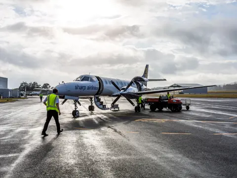 Ground crew in high visibility vests working around a Sharp Airlines propeller aircraft on a wet tarmac, King Island Airport