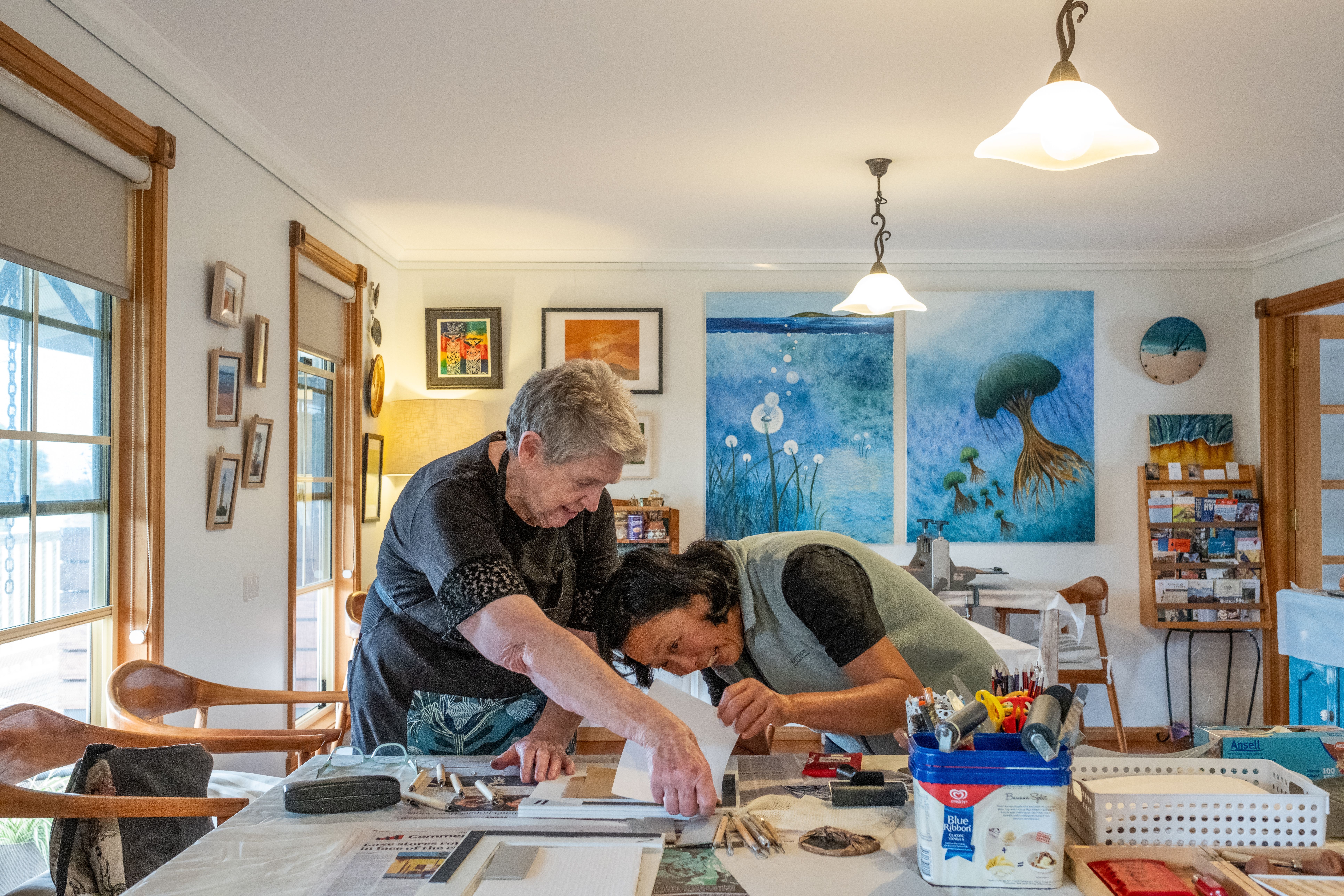 Two people work on a printmaking project at a table surrounded by art supplies and paintings at Beaumaris Beach Guesthouse