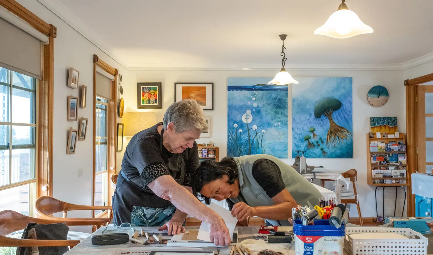 Two people work on a printmaking project at a table surrounded by art supplies and paintings at Beaumaris Beach Guesthouse