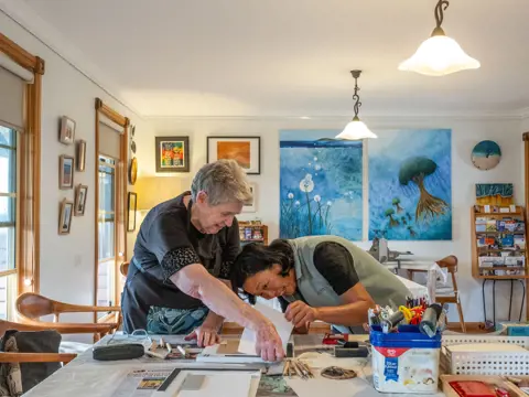 Two people work on a printmaking project at a table surrounded by art supplies and paintings at Beaumaris Beach Guesthouse