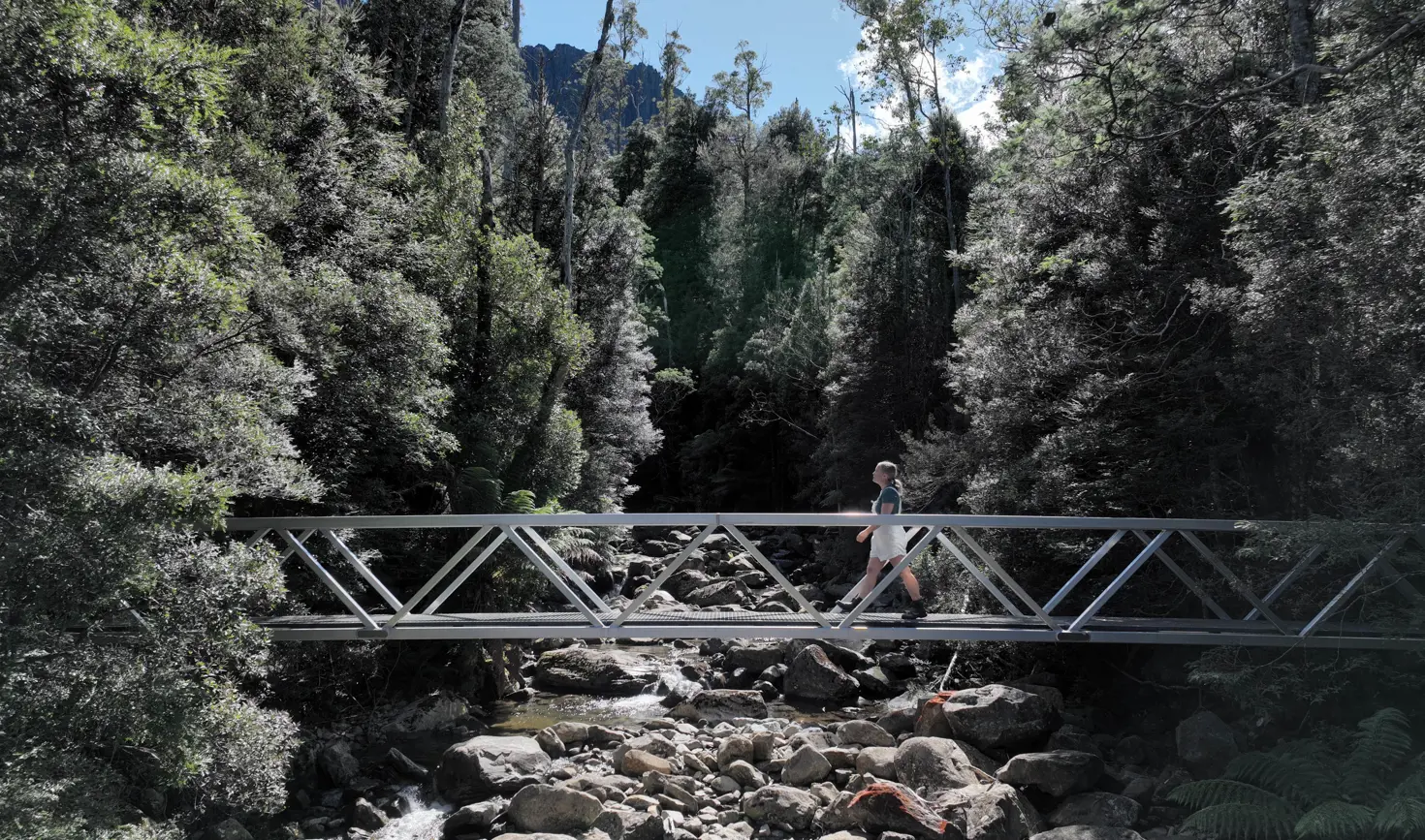 A walker crosses a metal footbridge over a rocky creek on the Smoko Falls Track, Meander Forest Reserve