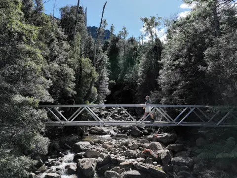 A walker crosses a metal footbridge over a rocky creek on the Smoko Falls Track, Meander Forest Reserve