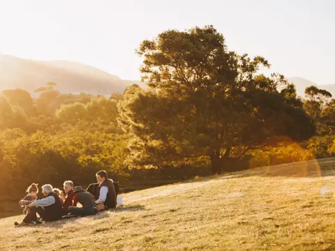 A group sits together on a grassy hillside in warm golden light with hills and trees behind at Coaldale Walnuts, Richmond