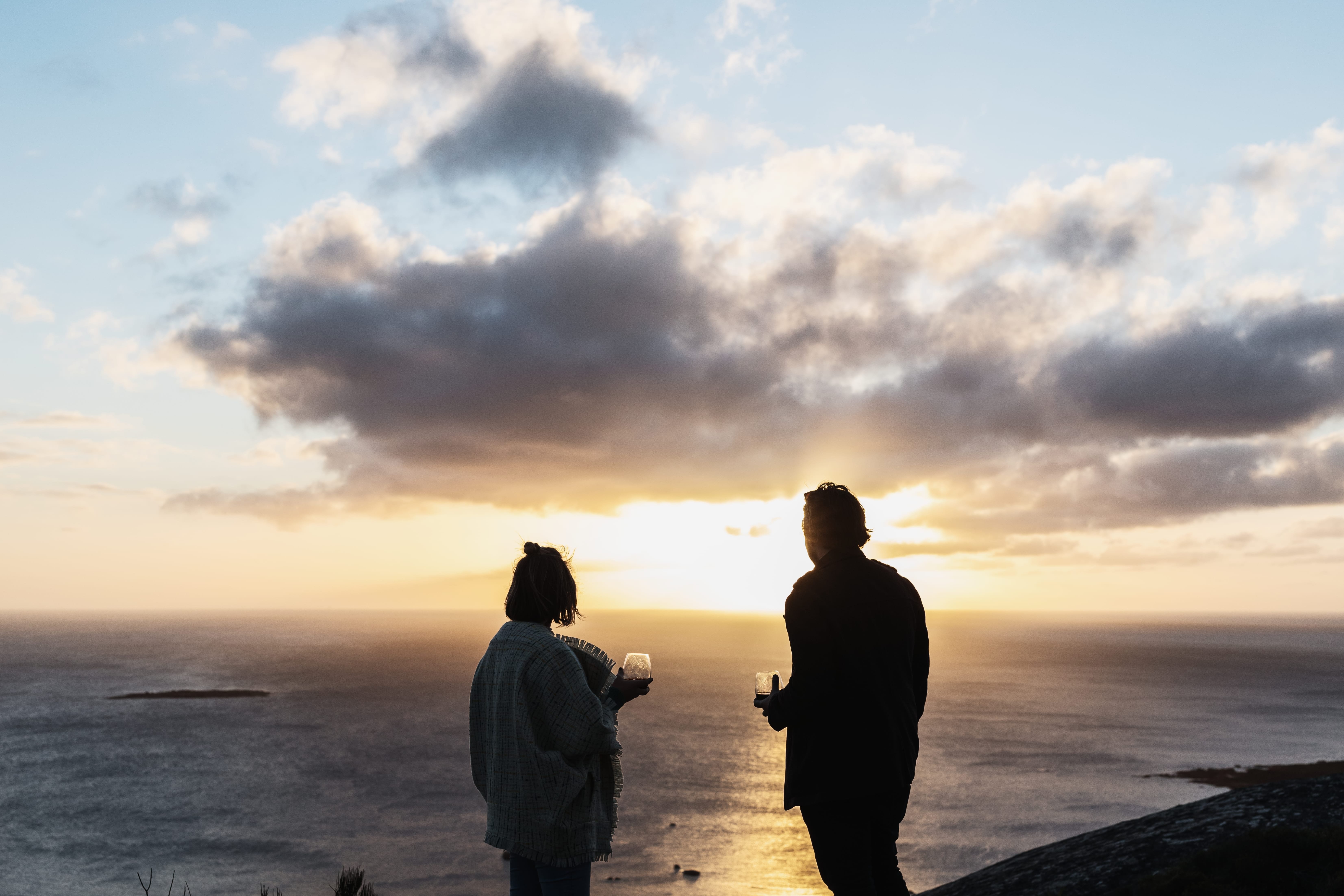Two people holding wine glasses look out over the sea at sunset from a clifftop on Flinders Island