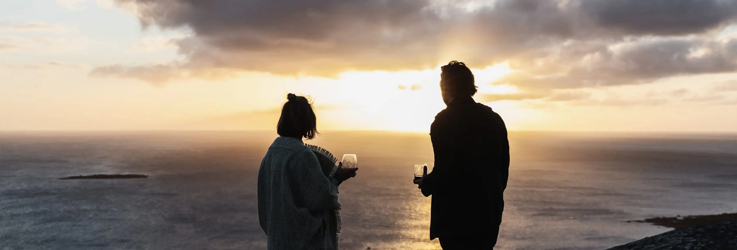 Two people holding wine glasses look out over the sea at sunset from a clifftop on Flinders Island