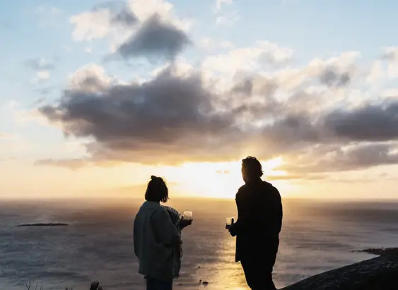 Two people holding wine glasses look out over the sea at sunset from a clifftop on Flinders Island