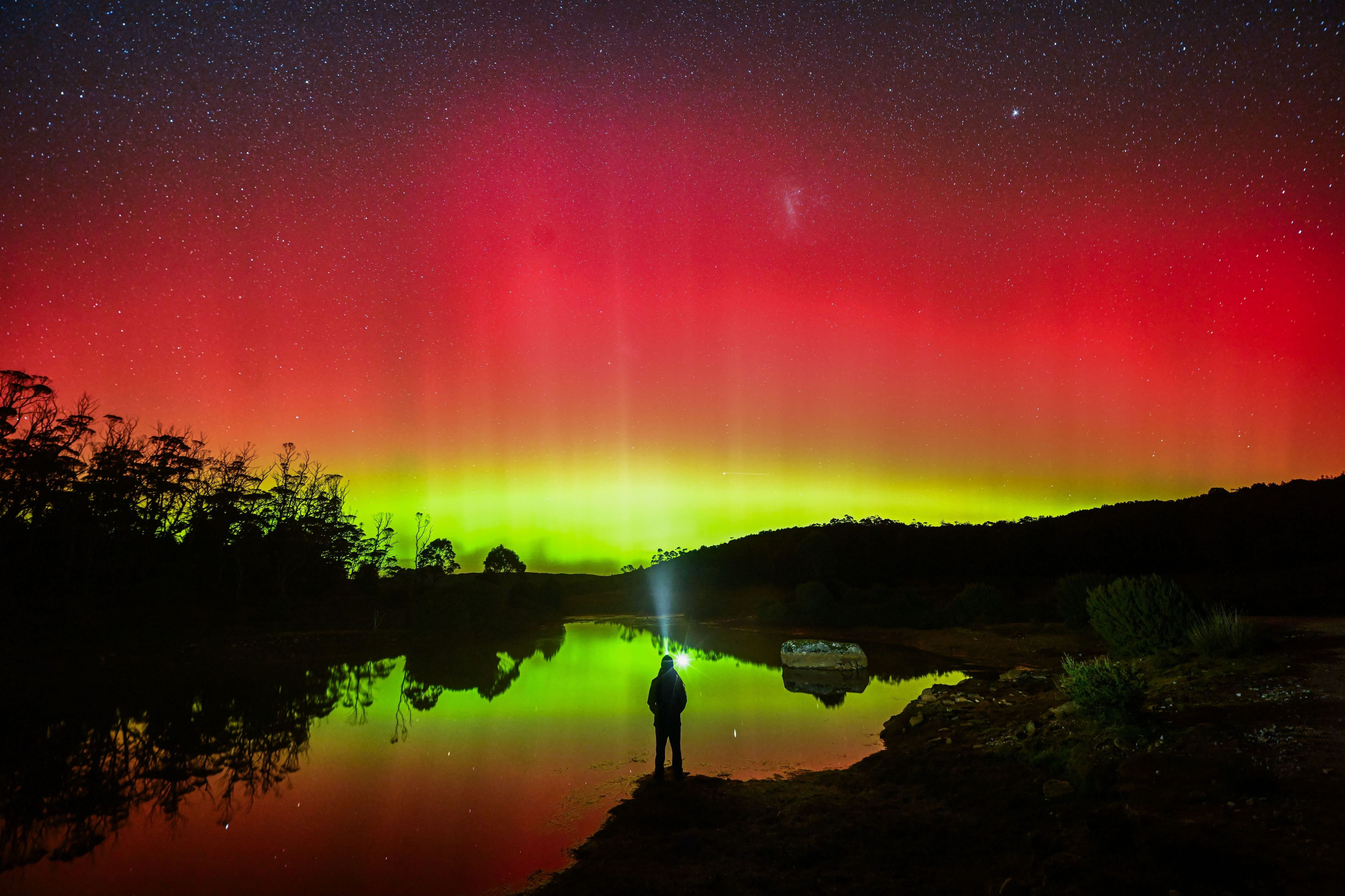 Person silhouetted by a lake watching the red and green Aurora Australis reflected in the water under a starry sky