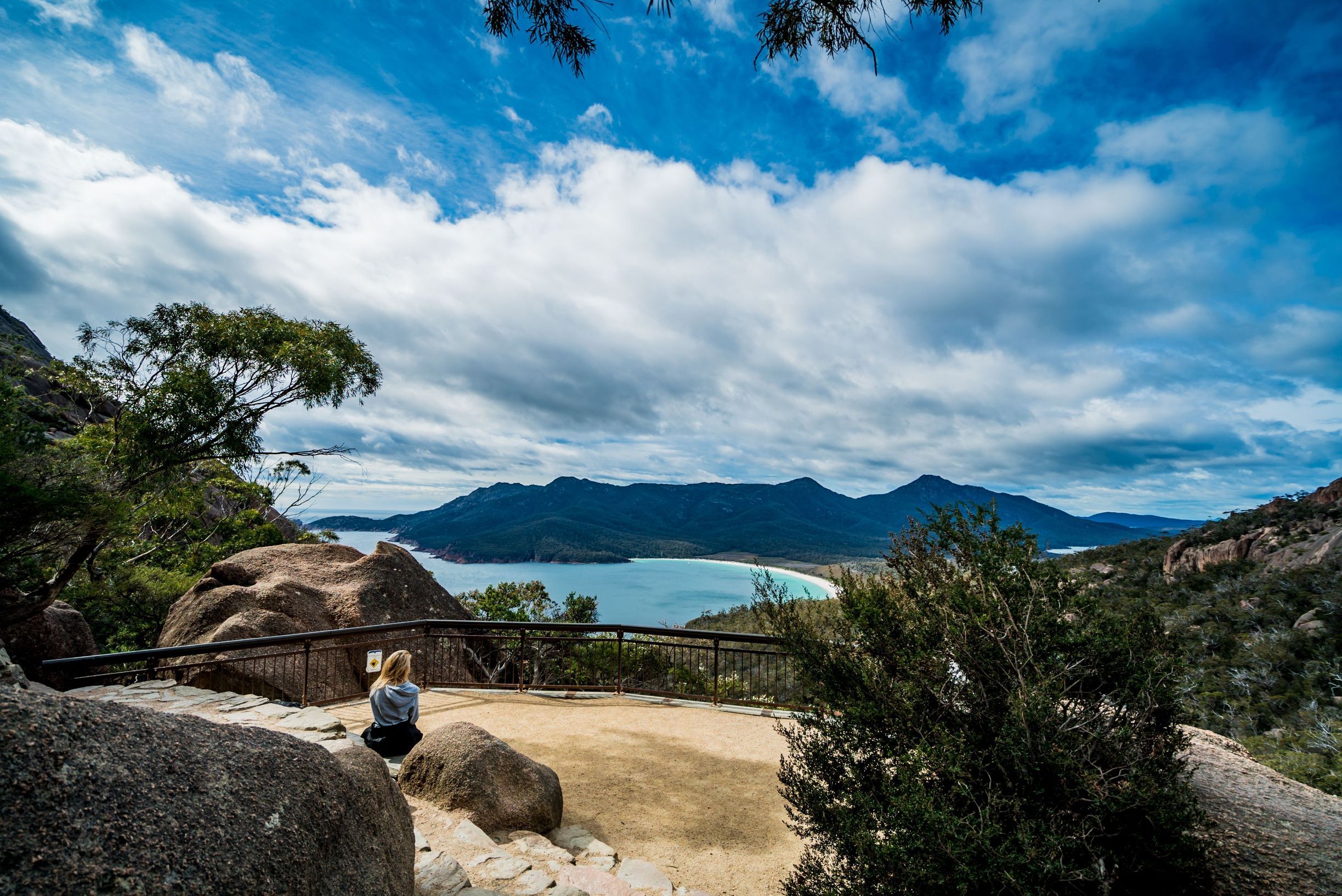 A person sits at the Wineglass Bay Lookout platform overlooking the curved beach below, Freycinet National Park