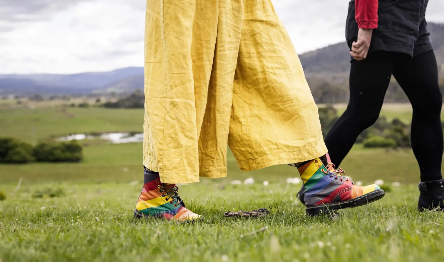 Two people walking on green grass at Twamley Farm, Buckland, one wearing rainbow boots and yellow trousers