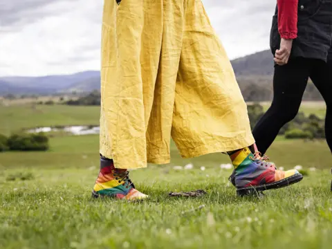 Two people walking on green grass at Twamley Farm, Buckland, one wearing rainbow boots and yellow trousers