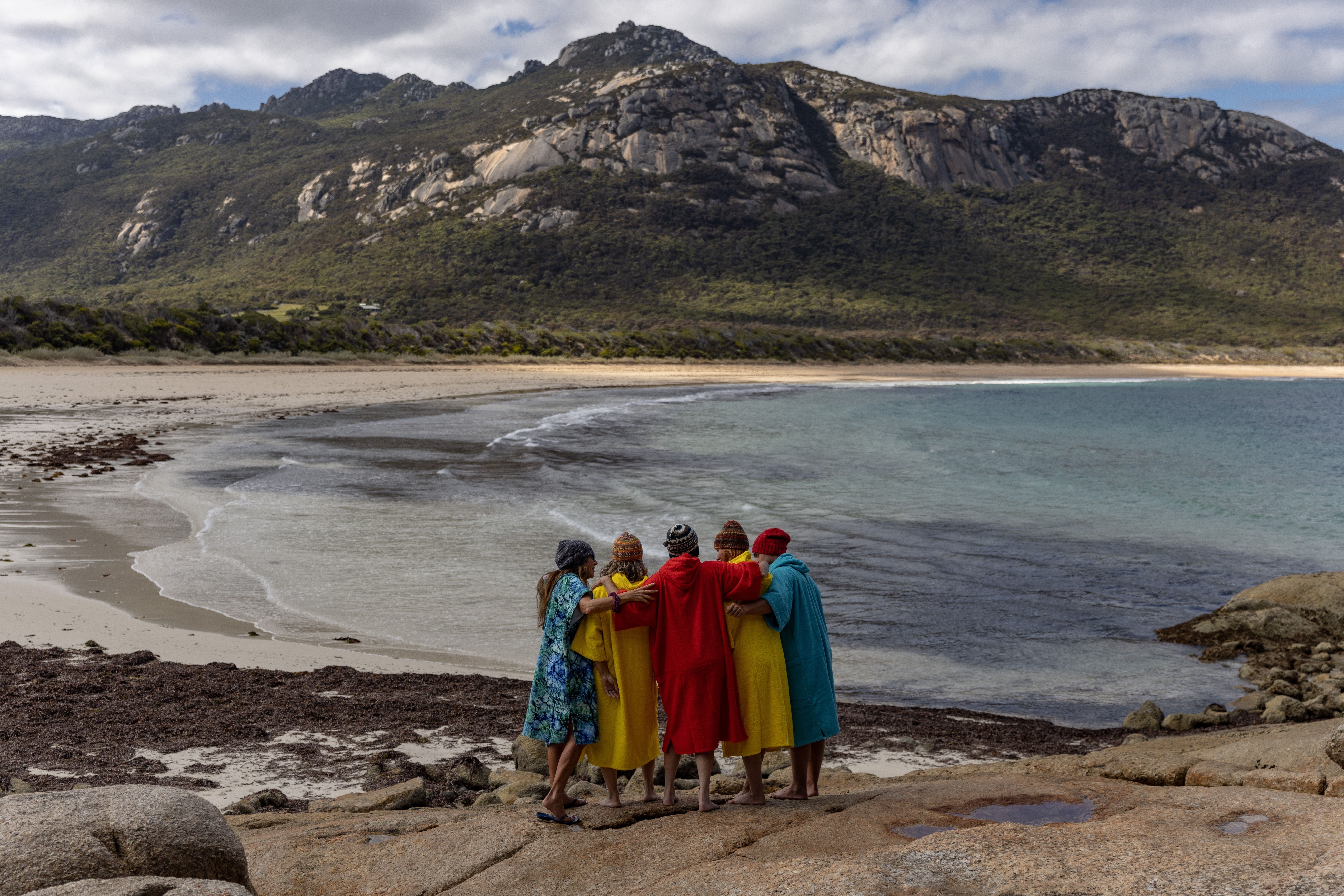 A group in colourful robes stand arm in arm on rocks overlooking a beach and mountains at FARAWAY on Flinders Island