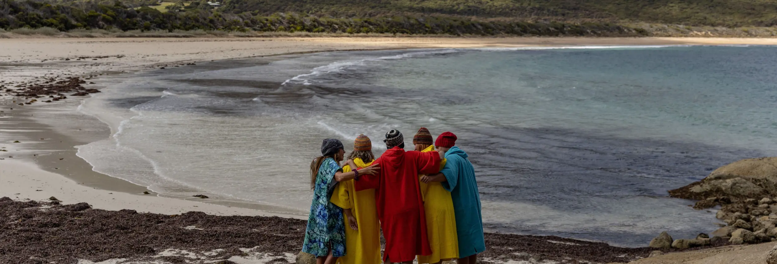 A group in colourful robes stand arm in arm on rocks overlooking a beach and mountains at FARAWAY on Flinders Island