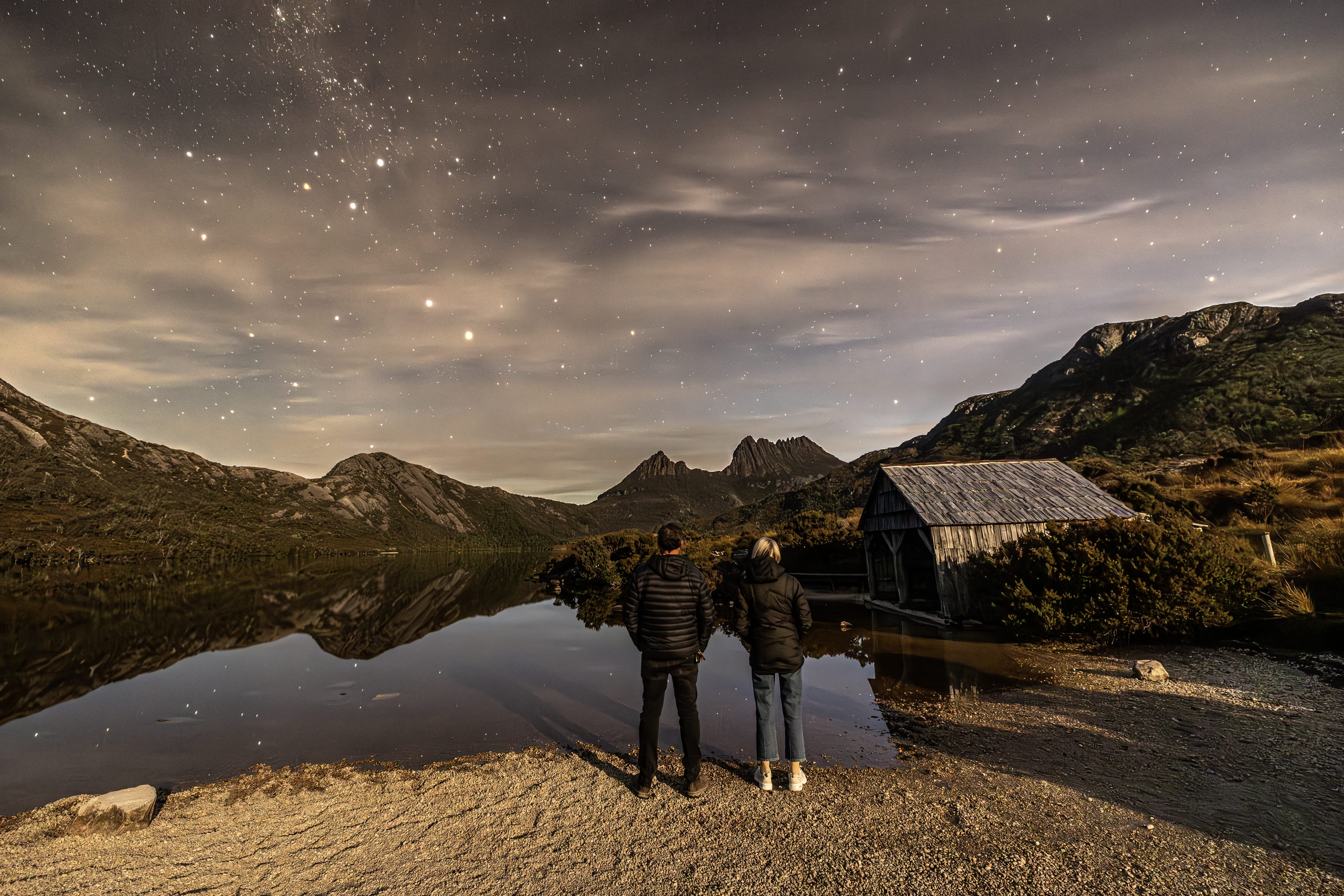 Two people stand at the edge of Dove Lake looking toward Cradle Mountain and a wooden boatshed in warm evening light