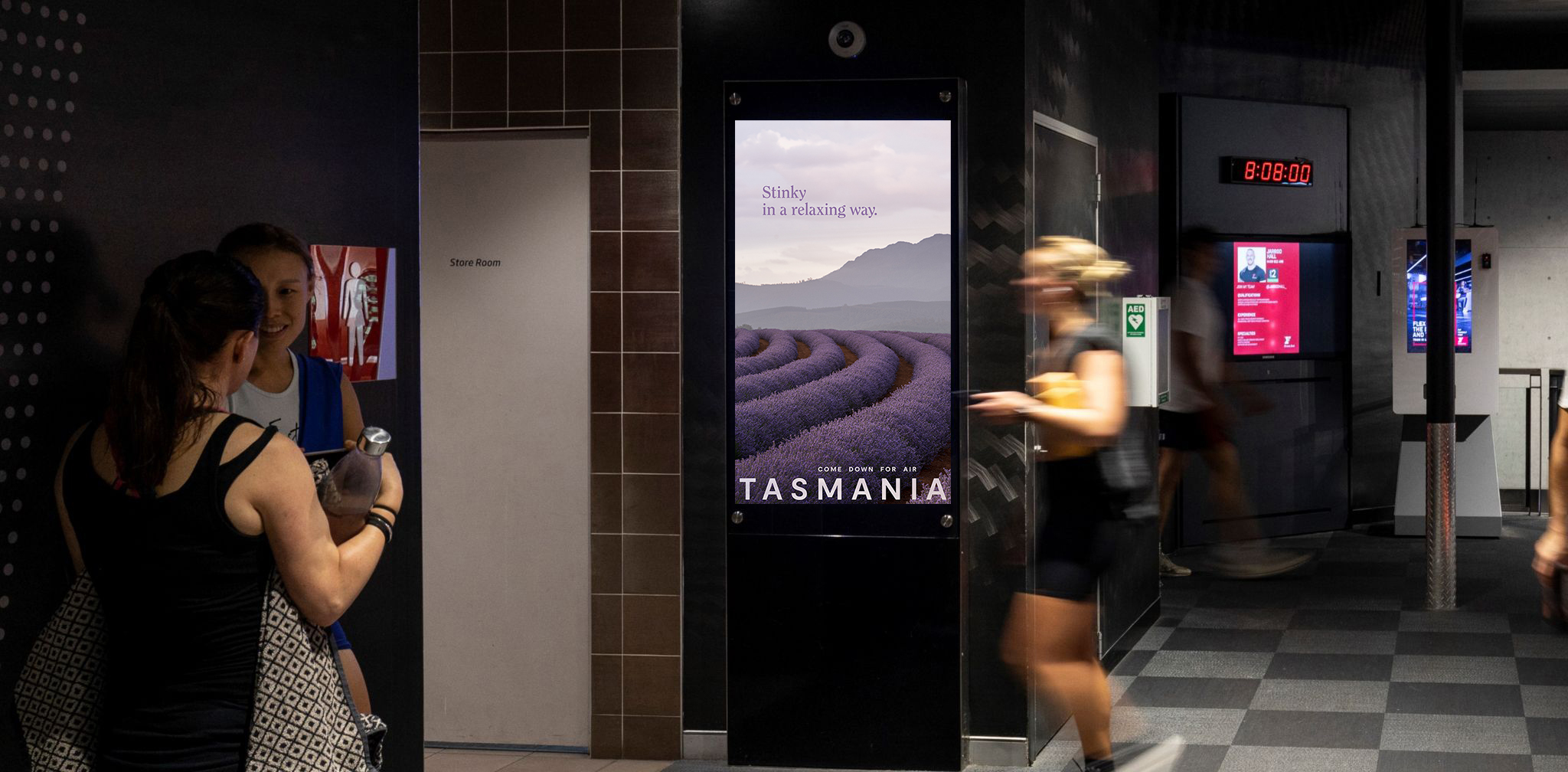 A Tourism Tasmania Come Down For Air campaign poster featuring lavender fields displayed on a digital screen in a gym