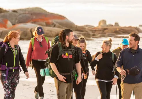 A Pakana / Palawa guide leads walkers along a sandy beach with orange lichen covered boulders, wukalina Walk