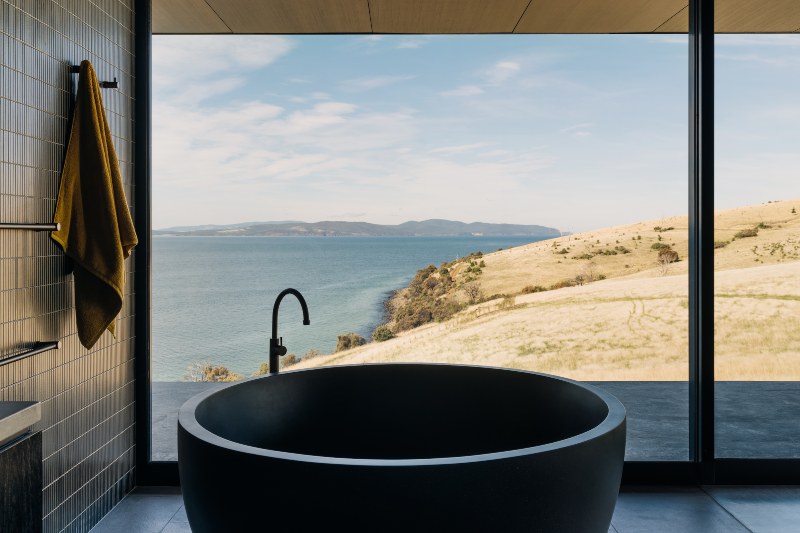 A deep, circular, dark stone bath has a perfect view through large windows down a grass hill to the water's edge.