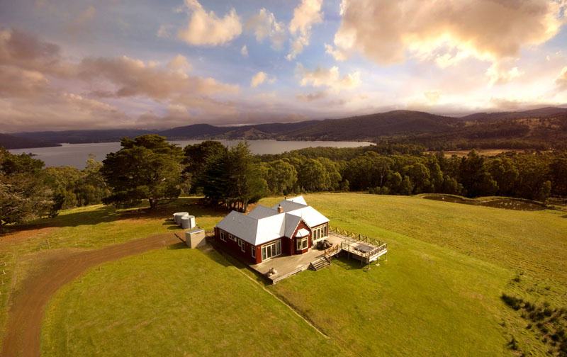 A large house sits on top of a green grass hill surrounded by forest, with a river and mountain in the background