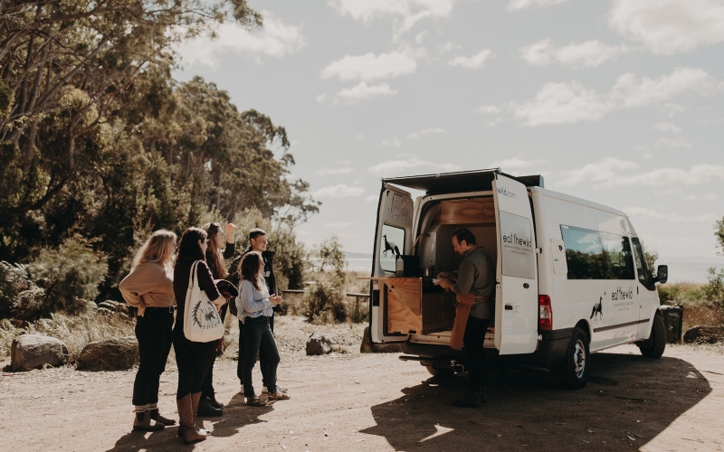 A group of people gather around a large van carrying a portable kitchen setup, parked in a picturesque site next to the beach.