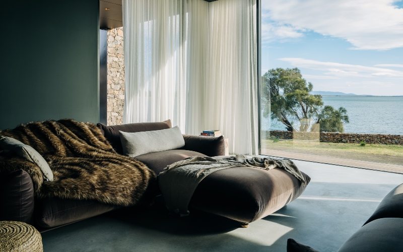 A stylish bedroom with large floor to ceiling windows looks out across the water on a sunny day.