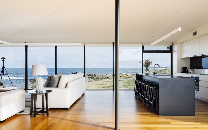A photograph of a modern interior with polished floor boards and floor to ceiling windows looks out over a beach
