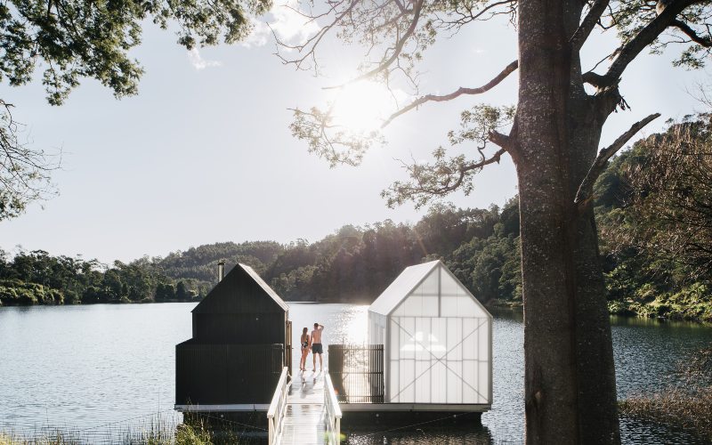 Two undercover saunas are at the end of a pier overlooking a lake on a slear sunny day. A man and woman enjoy the view.