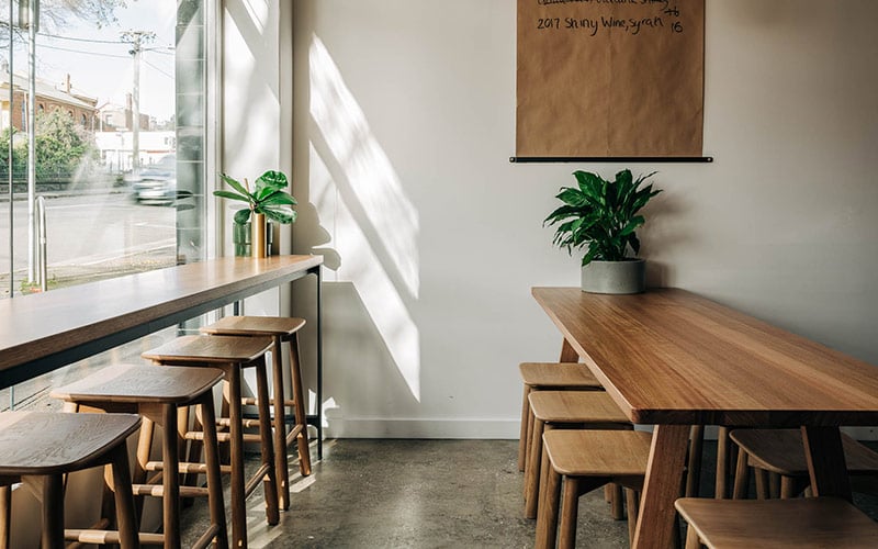 A bar with polished concrete floors and large windows is furnished with mid-century wooden benches and stools and green indoor plants 