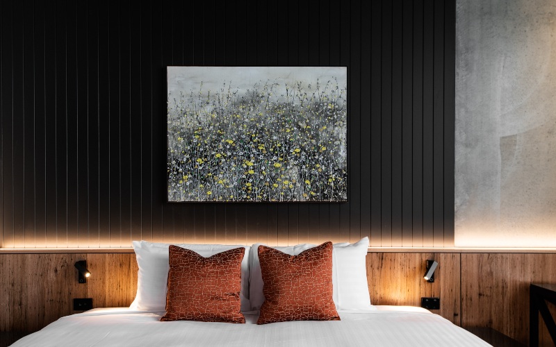 A contemporary painting of a flowers in a field is hung above a white sheeted bed with ochre coloured pillows in this photograph of a hotel room at The Verge