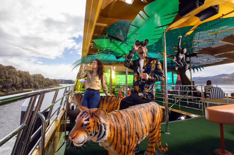 A young couple stand on the decks of the Mona Roma next to a plastic tiger, looking out across the water, sipping wine.