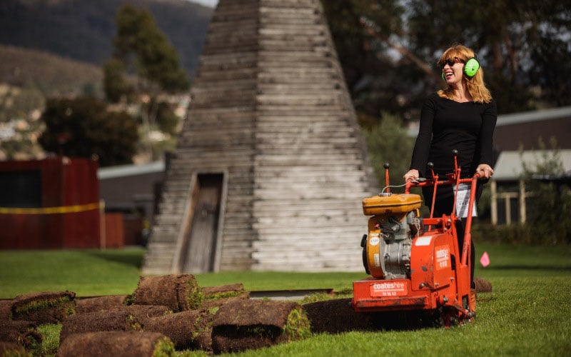A young woman smiling and wearing safety gear is pushing a motorised turf removing machine 