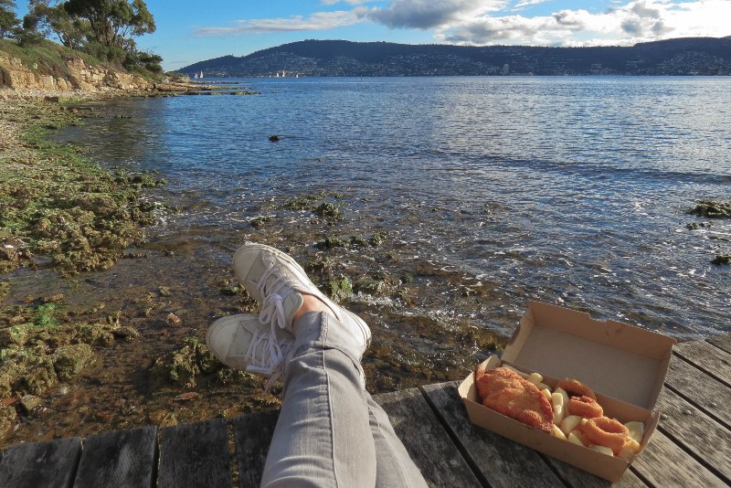 A woman sits on a wooden pier with fish and chips and looks out across the water. 