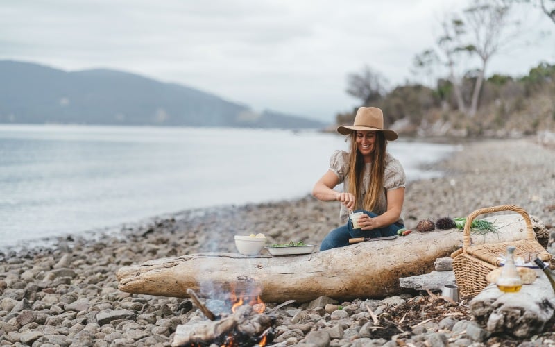 A woman sitting on a cobble-stone beach, cooking over  an open flame 