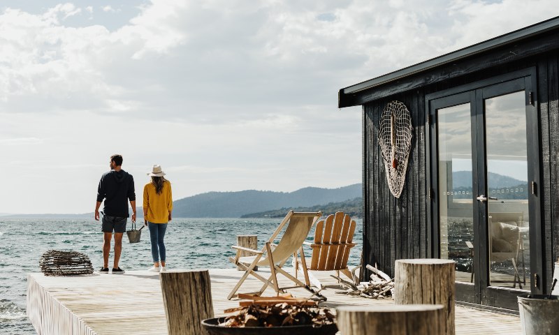 A man and a woman stand on the pier with an open fire pit and seating, in front of a luxury cabin on Satellite Island