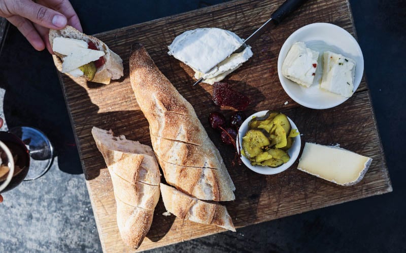 A loaf of sourdough bread, cheese and pickles are arranged on a wooden board 
