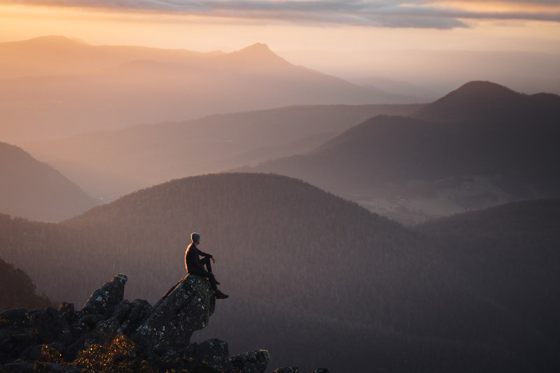 A man sits on a rocky outcrop looking across the valleys below kunanyi//Mt Wellington.