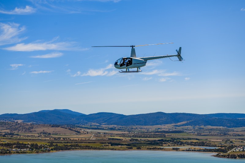 A 4 seater helicopter hovers over the blue waters of a bay on a sunny day.