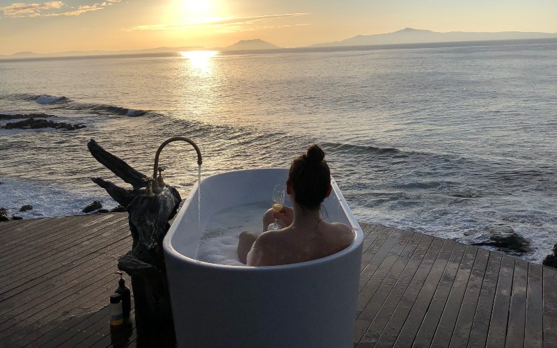 A lady sips at wine in an outdoor bath looking out over the ocean