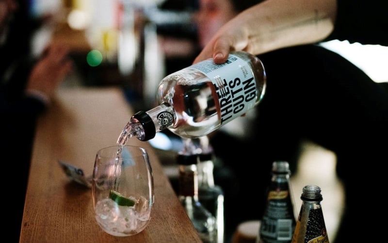 Gin is pured carefully over ice and lime in a stemless wine glass sitting on the bar counter.