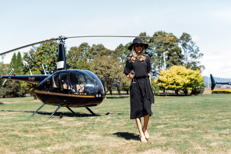 A woman in a dark dress and wide brimmed hat walks across the lawns in front of a small, 4 seter helicopter.