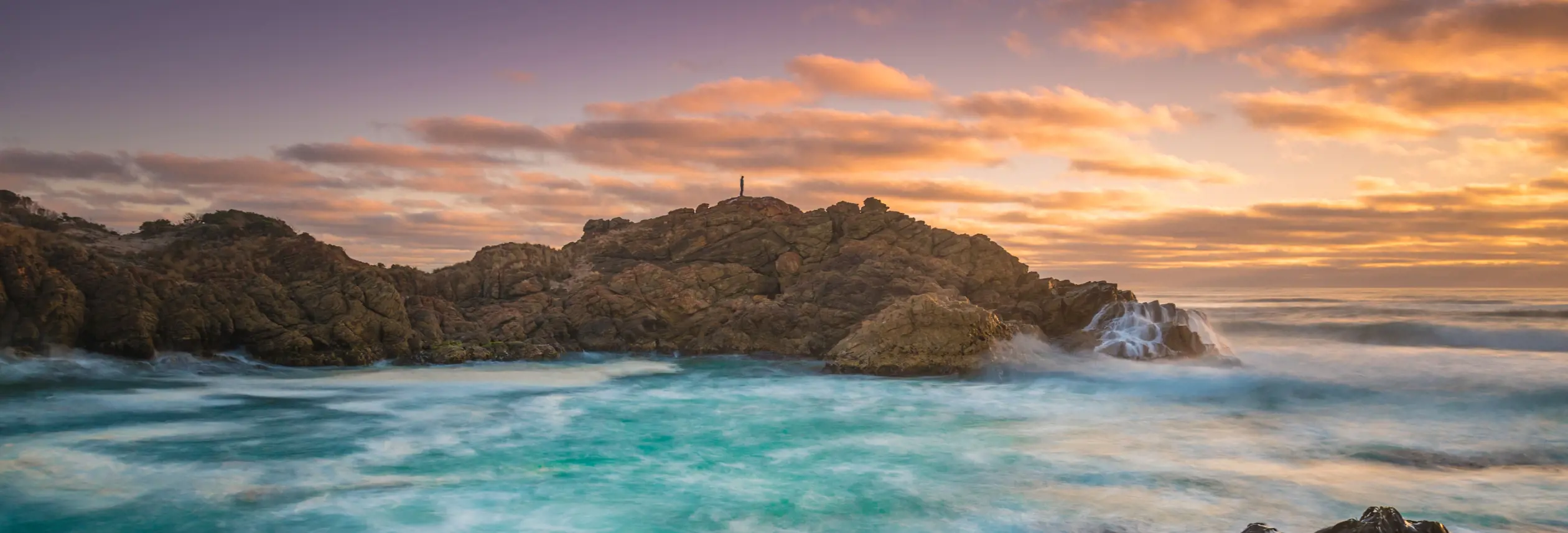A person silhouetted on a rocky headland with waves breaking into a turquoise bay at Trial Harbour, Tasmania's West Coast